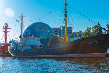museum ship SRT-129, moored at the embankment next to the Planetarium of the Museum of the World Ocean in Kaliningrad. It is an old fishing trawler with a black hull and yellow masts, carefully restored for museum purposes. In the background is the spectacular globe-shaped planetarium ball and modern buildings. To the right are reflections of sunlight on the water. To the left of the ship, a bright red lighthouse ship is partially visible. The scene is full of marine and scientific symbolism, and the day is clear and sunny.の写真素材