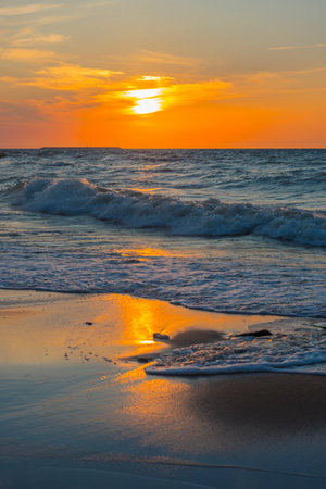 a picturesque sunset over the Baltic Sea. The sun slowly sinks below the horizon, painting the sky and water in rich shades of orange, yellow and blue. The waves gently roll onto the sandy shore, reflecting the golden light. In the foreground are wet sand covered with surf foam and wooden extending into the water. The atmosphere of calm, solitude and natural harmony makes the scene ideal for illustrating themes: relaxation, nature, meditation, travel.の写真素材
