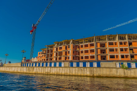 the process of active construction of a residential building on the river bank in Kaliningrad. In the foreground is water reflecting the construction site. The red brick building is under construction: there are no windows, concrete structures and wooden supports on the roof are visible. Tower cranes rise to the right and left. Everything is enclosed by a metal fence with blue inserts. The scene is illuminated by warm evening light, emphasizing the texture of the brick and creating a sense of movement and development.の写真素材
