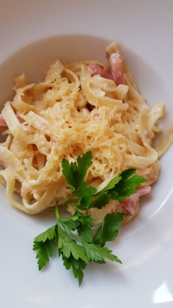 A mouth-watering serving of pasta in a creamy sauce, served in a deep white bowl. Fettuccine or tagliatelle-type pasta is topped with a creamy cheese sauce and sprinkled with grated cheese (perhaps Parmesan). The dish also includes bits of ham or bacon. A fresh sprig of parsley is added to the top, adding a bright green accent and a finished look. The presentation is neat and restaurant-quality.の写真素材