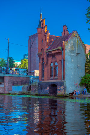 a picturesque red brick Gothic building standing right on the river bank in Kaliningrad. The corner turret with a high spire and decorative battlements give the building the appearance of a small castle or watchtower. The building can be recognized as one of the old water towers or security structures typical of KÃ¶nigsberg architecture. In the foreground is a water surface with a reflection of the building, and on the right are people relaxing with a dog. In the background are modern and historical brick houses.の写真素材