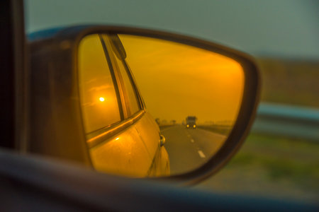 moment on a car trip - a view through the side rearview mirror of a car. The reflection of the mirror shows the setting sun, painting everything in a warm golden-orange tone. The horizon line, cars on the road and part of the car body create an atmosphere of coziness, the road and slight nostalgia. The effect of the warm filter gives the frame a cinematic and philosophical mood â as if looking back into the past.の写真素材