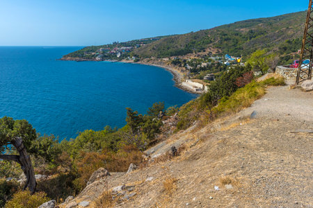 a magnificent view of the Crimean coast. From above, a panorama of the curved coastline, covered with sparse vegetation, rocks and junipers, opens up. In the distance, a small town or village with white houses spread out along the slope of a hill is visible. The Black Sea is deep blue, calm, with slight glare from the sun. On the horizon, closer to the center of the frame, an old sea platform or pier is noticeable. This place conveys an atmosphere of privacy, tranquility and southern warmth.の写真素材