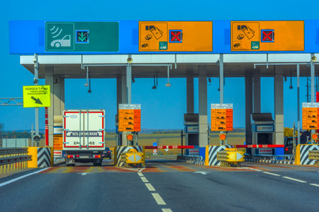 toll booth on the M4 Don toll road in Russia. In the foreground, a truck passes through a barrier in a special lane with a green arrow, intended for transponders (automated payment system). Above each lane, there are electronic boards with payment methods (cash, card, transponder) and accessibility information. The infrastructure is clearly organized: road signs, fences, CCTV cameras, and safety signs. In the background, there is an open rural landscape with fields and trees on the horizon.の写真素材