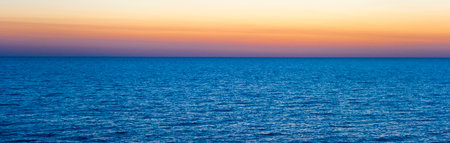 A wide panorama of the sea surface at sunset. The deep blue of the water contrasts with the soft pastel shades of the sky - from bright orange to pink and lilac. The calm surface of the sea emphasizes the peaceful atmosphere, creating an ideal background for themes related to nature, travel, meditation, freedom and relaxation.の写真素材