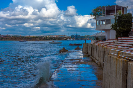 a picturesque view of Sevastopol Bay with a concrete embankment and the steps of an amphitheater facing the sea. Along the shore there is a white building with a terrace, and in the distance you can see ships and cranes of shipyards. The weather is sunny but cloudy, creating a striking contrast between the blue sky, sea and architecture. On the horizon you can see the cityscape of Sevastopol with its historical and industrial sites.の写真素材