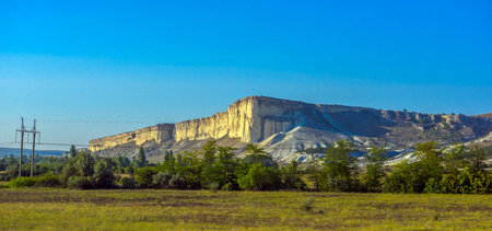 White Rock (Ak-Kaya) in Crimea is a monumental limestone formation with vertical cliffs, towering over a green valley. The rocky massif is flooded with sunlight, emphasizing the texture and light cream shade of the limestone. In the foreground are meadows and trees, creating a picturesque natural contrast with the majestic relief of the mountains. The sky is clear, bright blue, which adds a sense of space and grandeur to the frame.の写真素材