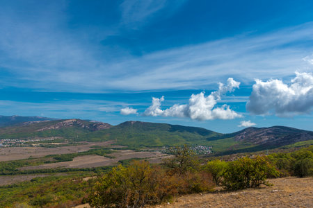 A picturesque view of a Crimean valley from a hill. In the foreground is a dry grassy plain with bushes and trees, the color of which hints at the onset of autumn. In the distance stretch green hills and mountains covered with forests, at the foot of which small villages are visible. The blue sky with light clouds emphasizes the space and natural beauty of the southern landscape. The scene is filled with a sense of calm, warmth and silence.の写真素材