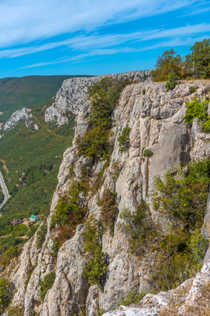 An impressive rocky cliff in the Crimean mountains overlooking a serpentine road that stretches along wooded slopes. In the foreground is a textured cliff surface covered in dry grass and moss. In the distance are mountain ranges, partly covered in greenery, under a bright blue sky with light clouds. Epic and picturesque landscape is perfect for themes of active recreation, mountaineering, hiking and natural power.の写真素材