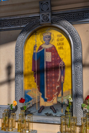 The photograph depicts an icon of the Holy Prince Vladimir, Equal to the Apostles, installed in a carved stone icon case on the territory of the New Chersonesos historical and cultural complex in Sevastopol. The holy prince is depicted in royal vestments with a cross and a sword, on a gold background, symbolizing holiness. Glass vases with live roses are installed in front of the icon, emphasizing the veneration and solemnity of the place. The Byzantine ornament of the frame emphasizes the sacred nature of the composition.の写真素材