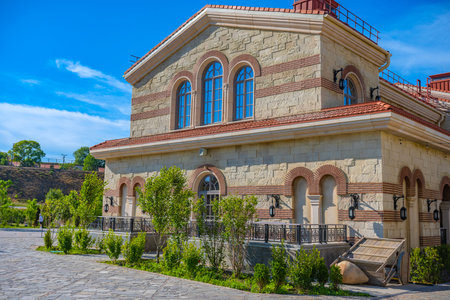 the building is in an architectural style that combines elements of the Byzantine and southern Russian traditions. Light stone and decorative brick inserts give the building expressiveness and a historical appearance. Windows with arched openings and black forged lanterns emphasize the style of the reconstructed cultural complex New Chersonesos in Sevastopol. In front of the building there are neat bushes, well-kept paths, and by the wall there are stylized elements of antiquity, including a cart and a ceramic vessel. The scene is filled with summer sunlight and the atmosphere of a southern resort town.の写真素材