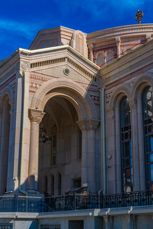 An architectural fragment of the building of the cultural and historical complex "New Chersonesos" in Sevastopol. In the foreground is a massive arch with decorative carvings and columns made of light stone. The architecture contains elements of the Byzantine and neoclassical styles: arched windows, geometric ornamentation, panels and carved capitals. Clear sunlight emphasizes the relief of the details. In the background is a bright blue sky, creating a visual contrast with the beige and pink palette of the facade. The building looks monumental, emphasizing the connection with the cultural heritage of Chersonesos.の写真素材