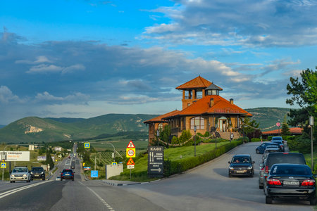 A modern cafe with a red tile roof against the backdrop of picturesque Crimean hills and green valleys. The building in the style of Mediterranean architecture rises on the side of the highway leading deep into the mountainous Crimea. In the foreground is a parking lot with cars and a sign with the establishment's menu. Soft evening light emphasizes the atmosphere of coziness and hospitality, creating an ideal scene for travelers.の写真素材