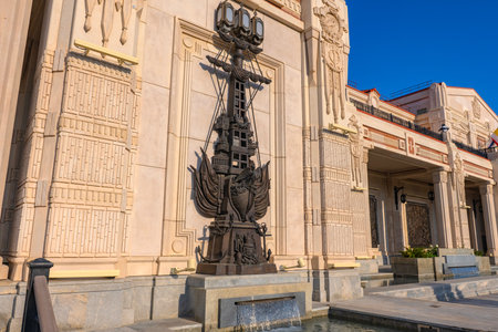 A modern neoclassical building with rich architectural decor, located in the New Chersonesus complex in Sevastopol. In the center of the frame is a massive bronze composition in the form of a stylized ship, decorated with sails and maritime symbols. The composition is installed on a light stone faÃ§ade, against which it stands out with its contrast and artistic elaboration. In front of the sculpture is a small fountain with falling water. The bright daytime sun emphasizes the texture of the materials, architectural details and creates a solemn, monumental impression.の写真素材