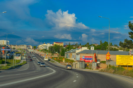 A city street in Crimea against the backdrop of a picturesque sky with powerful cumulus clouds. In the foreground, you can see a bus stop and a protected civil defense shelter â a concrete structure designed to provide emergency shelter during missile or drone attacks. Public transport is actively moving along the road, construction work and information boards are visible. Modern urban infrastructure is combined with security elements in a changing geopolitical situation.の写真素材