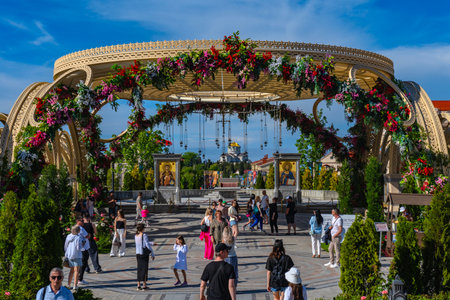 The photo was taken in the cultural and historical complex "New Chersonesus" in Sevastopol. In the foreground is an arched structure decorated with fresh flowers with hanging lamps, forming the entrance to the ceremonial site. Inside are icons, marble pedestals, a landscaped area and many visitors, including children and adults. In the distance, along the axis of the composition, is a majestic temple with a golden dome, symbolizing the spiritual axis of space. The atmosphere is bright, festive, with elements of traditional Orthodox culture and modern landscape design.の写真素材