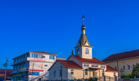 A bright sunny day in Crimea. In the foreground is a small Orthodox church with a golden dome and a cross, typical architecture with a red metal roof and white walls. Nearby is a typical building with shops and signs, including an auto parts store and a microfinance organization. The combination of a religious building and an everyday urban environment highlights the contrasts of modern life. In the background is a clear and rich blue sky, without a single cloud. The photo conveys the atmosphere of a southern town with elements of tradition and modern commerce.の写真素材
