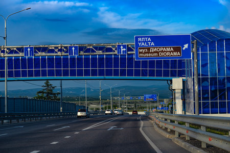 A modern highway in Crimea with directional road signs indicating the way to Yalta and the Diorama Museum. The panorama opens up to a view of the mountain landscape in the distance, a clear sky with light clouds and an architectural element in the form of blue glazing. The asphalt highway is multi-lane, cars move along it, emphasizing the active transport artery of the region. The infrastructure is built according to modern standards, with noise protection screens and good markings.の写真素材