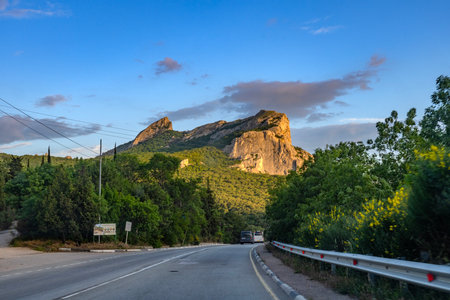 Landscape photo of the Crimean highway against the backdrop of a majestic mountain range. The central place is occupied by a picturesque rock, bathed in the soft light of the setting sun. The road goes into the distance, leading travelers through green hills and forests. Along the side of the road there is lush vegetation, bushes with yellow flowers and a road fence. Cars are moving in the distance, emphasizing the atmosphere of the road and freedom. An ideal illustration of Crimean nature and car travel.の写真素材