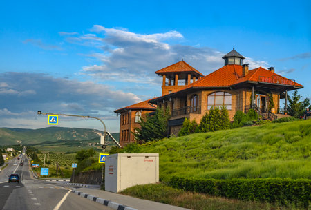 A Tuscan-style tiled-roof cafe on a picturesque hilltop by a highway in Crimea. In the foreground is a white concrete civil defense shelter, designed to protect against drone or missile attacks. It is built next to a busy road that leads through rolling green expanses. Mountains and vineyards can be seen in the distance, and the sky is adorned with soft clouds, creating a contrast between the peaceful nature and the anxious need for protection.の写真素材