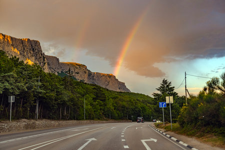 Picturesque landscape of the southern coast of Crimea at sunset. The asphalt road leads along a dense green forest to a rock massif, illuminated by the soft orange light of the setting sun. Two rainbows spread out over the mountains, one of which stands out especially brightly against the background of clouds. The unique combination of natural phenomena - sunset light, dramatic clouds and a double rainbow - creates an atmosphere of magic and peace. In the frame - a road with markings, cars, a pine forest and majestic rocks, typical of Crimea.の写真素材