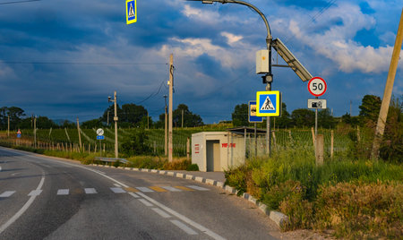 A photograph of a rural road in Crimea against the backdrop of a picturesque evening sky with dense clouds. Along the road is a bus stop and a modest concrete shelter designated as an air raid shelter. Above the road there are road signs: a pedestrian crossing, a speed limit of 50 km/h, cameras recording violations. In the distance are vineyards and trees, creating an atmosphere of southern solitude and uneasy calm. The combination of rural landscape, military infrastructure and civil security creates a contrasting and meaningful visual composition.の写真素材