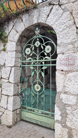 decorative metal gates, painted turquoise-green, with an elegant floral pattern. The gates are installed in an arched niche of a stone wall made of large limestone blocks. Behind the gates you can see an old stone staircase leading up. The metal surface is covered with patina and rust, which gives the image an atmosphere of antiquity and southern comfort. The photo was taken in the village of Simeiz, located on the southern coast of Crimea.の写真素材