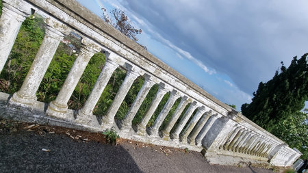 an old white stone balustrade with classical columns, partly covered with moss and traces of time. It is located on a cliff above a green valley, overlooking the sea and the horizon, above which a rainbow is visible. At the bottom of the railing there are cigarette butts and garbage, which creates a contrast between the noble architecture of the past and the indifference of the present. Dark clouds hang over the railing, and in the distance you can see the blue sky. The atmosphere of the photo is a mixture of desolation, beauty and deep silence.の写真素材