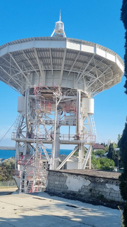 close-up of a massive radio telescope on the coast of Crimea. The metal structure with many engineering elements and ladders emphasizes the complexity and scale of the device. In the background, you can see the blue sea, green tree crowns and roofs of buildings, which creates a visual contrast between the high-tech structure and the natural landscape. This photo reflects the harmonious proximity of science, technology and nature, emphasizing the strategic importance of Crimea as a scientific and research center.の写真素材