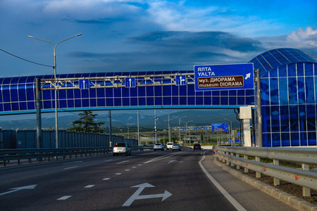 A modern highway in Crimea with directional road signs indicating the way to Yalta and the Diorama Museum. The panorama opens up to a view of the mountain landscape in the distance, a clear sky with light clouds and an architectural element in the form of blue glazing. The asphalt highway is multi-lane, cars move along it, emphasizing the active transport artery of the region. The infrastructure is built according to modern standards, with noise protection screens and good markings.の写真素材