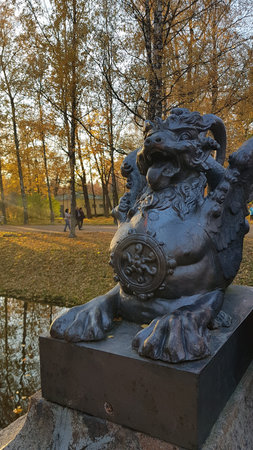 A bronze sculpture of a mythical winged lion with an open mouth, protruding tongue and an expressive grimace, installed on a stone pedestal in the park of the city of Pushkin (former Tsarskoye Selo). On the chest of the figure is a decorative medallion with a relief ornament. The statue is located on the bank of a park canal, the water of which reflects autumn trees. Around it are golden foliage covering the ground and paths. In the distance, people walking and small buildings are visible. The warm light of the setting sun gives the scene an atmosphere of coziness and tranquility, emphasizing the rich texture of the metal and the autumn palette of the parkの写真素材