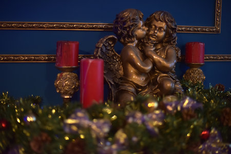 decorative composition with a bronze sculpture of two angels sitting together and tenderly embracing. One angel kisses the other on the cheek, creating an atmosphere of warmth, tenderness and peace. Around are red candles on carved golden candlesticks, as well as festive decor with fir branches, ribbons and shiny ornaments. The background is made in a rich blue color with gilded frames, which gives the scene sophistication and solemnity. The composition is ideal for Christmas or New Year themes, as well as for illustrating romantic and religious stories.の写真素材