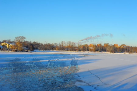 winter landscape with a frozen river or lake. In the foreground, the ice is cracked and has areas of open water reflecting the blue sky. The surface is covered with snow and fine ice patterns. On the horizon, there are bare trees, buildings and a factory chimney emitting smoke, creating the atmosphere of a winter industrial city. The low sun paints the trees and buildings in golden-orange tones, emphasizing the contrast between the cold shades of the ice and the warmth of the sunlight. The clear blue sky enhances the feeling of a frosty day.の写真素材