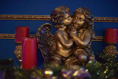 decorative composition with a bronze sculpture of two angels in a tender embrace. One of the angels with wings gently cuddles up to the other, creating an atmosphere of warmth, care and spiritual harmony. The sculpture is located against the background of a dark blue wall with a gold ornament, which gives the image nobility and sophistication. On the sides of the composition are red candles on carved candlesticks, decorated with gold details. At the bottom of the frame, New Year's tinsel is visible, adding a festive mood. The atmosphere of the photo combines Christmas symbolism, coziness and a sacred feeling of peace.の写真素材