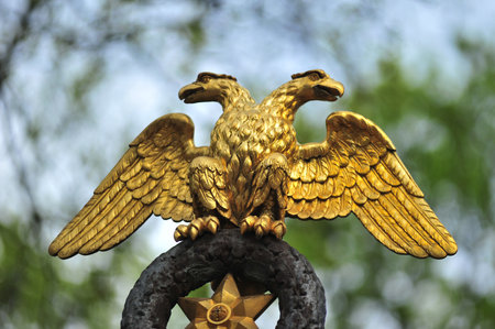 a golden double-headed eagle â a symbol of power, strength and grandeur. The sculpture is detailed: each feather is carefully worked out, the wings are spread out to the sides, the bird's heads are looking in opposite directions. The eagle is mounted on a wreath with a golden star, symbolizing solemnity and significance. In the background, there are softly blurred green tree crowns and a blue sky, which emphasizes the brightness and shine of gold. The image reflects historical and cultural symbolism, used in heraldry, architecture and official attributes.の写真素材