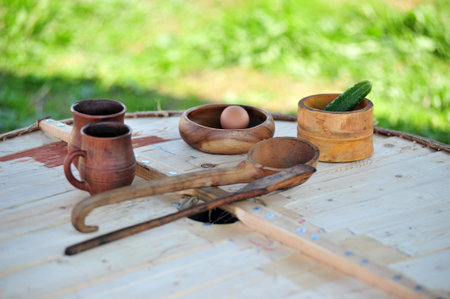 wooden table with traditional dishes and household items in rustic style. On the surface there are two wooden cups with handles, wooden bowls, one of which contains a chicken egg, the other - a fresh cucumber. There are also large wooden spoons and ladles, hand-carved. All objects have the characteristic texture and shades of natural wood. The background is green grass, slightly blurred, which emphasizes the rustic atmosphere and authenticity of the scene. The picture symbolizes simplicity, tradition and eco-friendliness of natural utensils.の写真素材