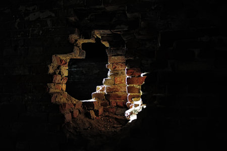 An atmospheric photograph of an old brick wall with a large hole through which light shines. The...