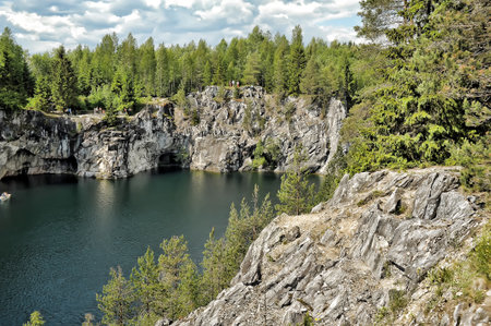 Marble Canyon in the Ruskeala Mountain Park (Karelia). The water is a deep emerald shade and is framed by steep gray cliffs with caves and cracks that go straight into the lake. Dense coniferous and deciduous forests grow along the banks, creating a beautiful contrast with the stone walls. Tourists can be seen admiring the panoramic view on the upper platforms of the canyon. On the right side of the frame is a rocky cliff with growing pines and firs, going down to the water. The atmosphere of the photograph conveys the beauty of northern nature, the tranquility and grandeur of a natural monument popular with tourists.の写真素材