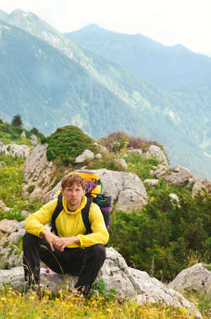 Young Man Traveller with backpack sitting on stones with Mountains on Backgroundの写真素材