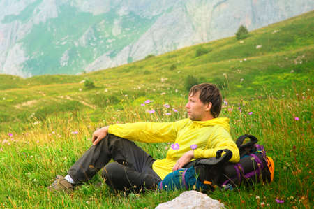 Young Man Traveller with backpack sitting on grass with flowers relaxing with Mountains on Backgroundの写真素材