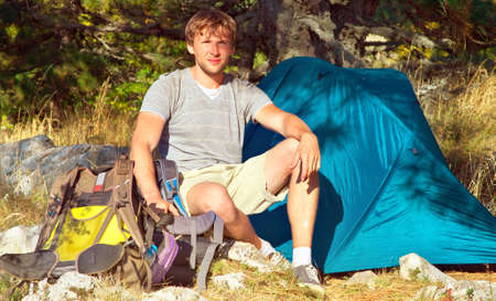 Young Man with Smiling Face Hiker sitting with backpack and Tent Camping Outdoor on Grass with forest nature on background Travel and Healthy Lifestyleの写真素材