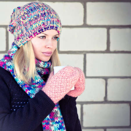 Young Woman with cofee cup Winter time wearing knitted sweater, hat and scarf with mittens Lifestyle concept trendy colors with white brick wall on backgroundの写真素材