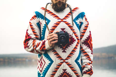 Young Man bearded with retro photo camera Fashion Travel Lifestyle wearing knitted sweater clothing outdoor foggy nature on backgroundの写真素材