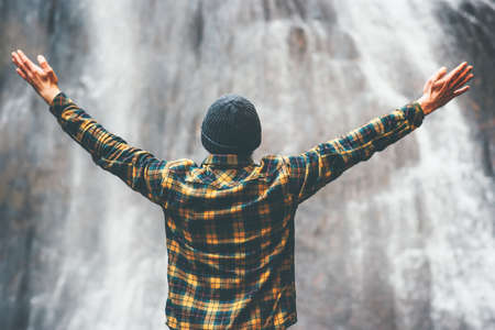 Man enjoying waterfall raised hands Travel Lifestyle adventure concept vacations into the wild wearing cozy shirt and hatの写真素材