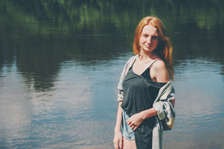 Pretty girl walking at river Lifestyle summer vacations harmony with nature concept. Young woman red hair wearing jeans shorts and top casual fashion clothingの写真素材