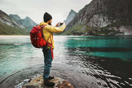 Man with backpack taking photo by smartphone walking on Norway beach Travel lifestyle wanderlust concept adventure outdoor summer vacations  の写真素材