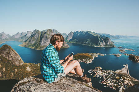 Man traveler using smartphone sitting on mountains top Traveling lifestyle concept adventure outdoor Reinebringen mountain aerial view Lofoten islands summer vacations in Norwayの写真素材