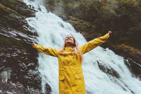 Young woman traveling at waterfall emotional raised hands adventure healthy lifestyle harmony with nature concept girl wearing yellow raincoat walking aloneの写真素材