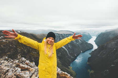 Woman traveling in mountains happy smiling raised hands healthy lifestyle adventure vacations aerial Norway fjord view girl wearing yellow raincoat の写真素材
