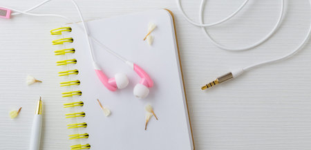 Pink earphones on notebook with pen and flower on white desk, top view
